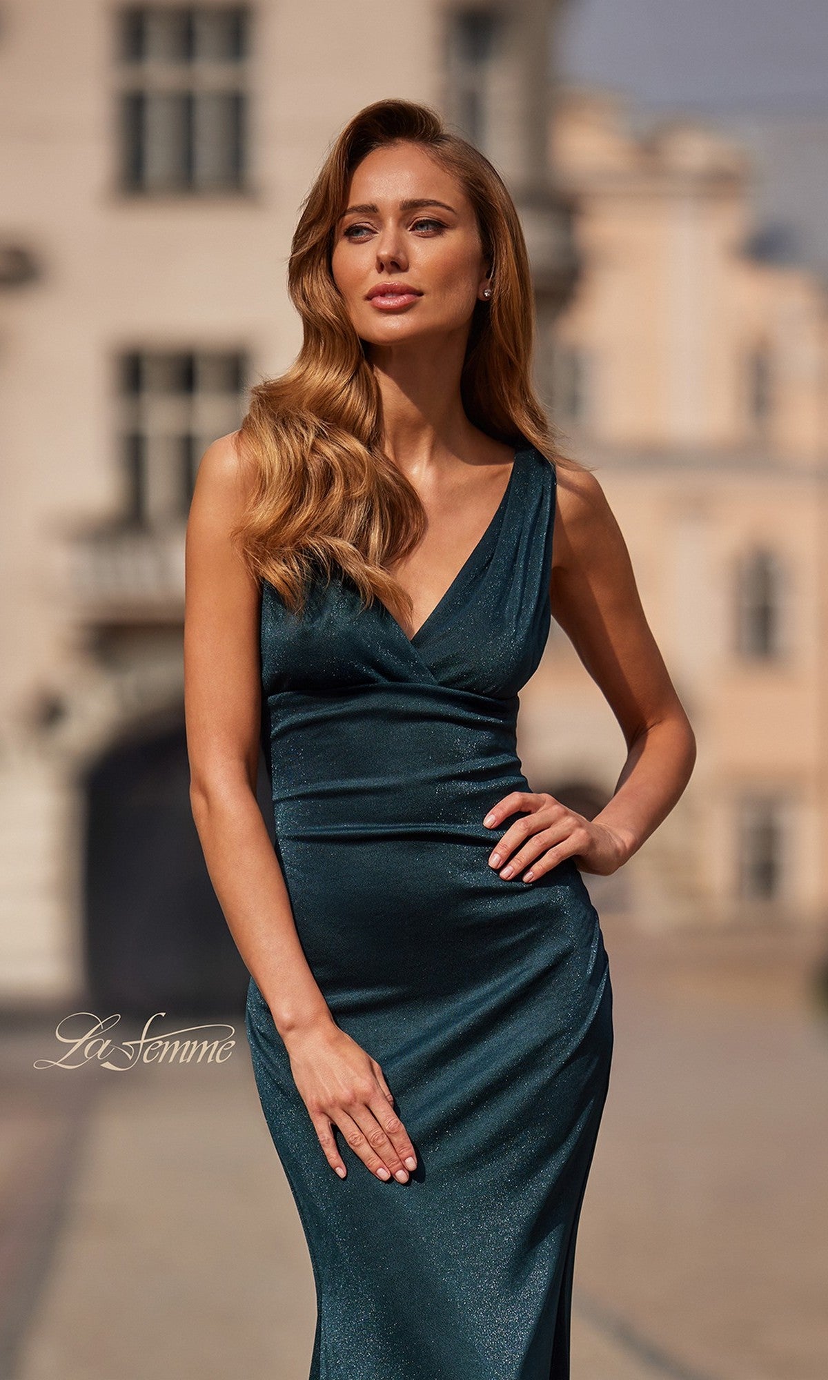 A woman with long wavy hair wears the La Femme 33425, a fitted, sleeveless dark green metallic jersey prom dress with a v-neckline, standing outdoors before blurred buildings.